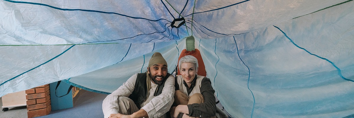 Two dancers sit side by side on the floor of a children's library, huddled up underneath a huge blue parachute, which is domed over their heads to look like a cave or a den.