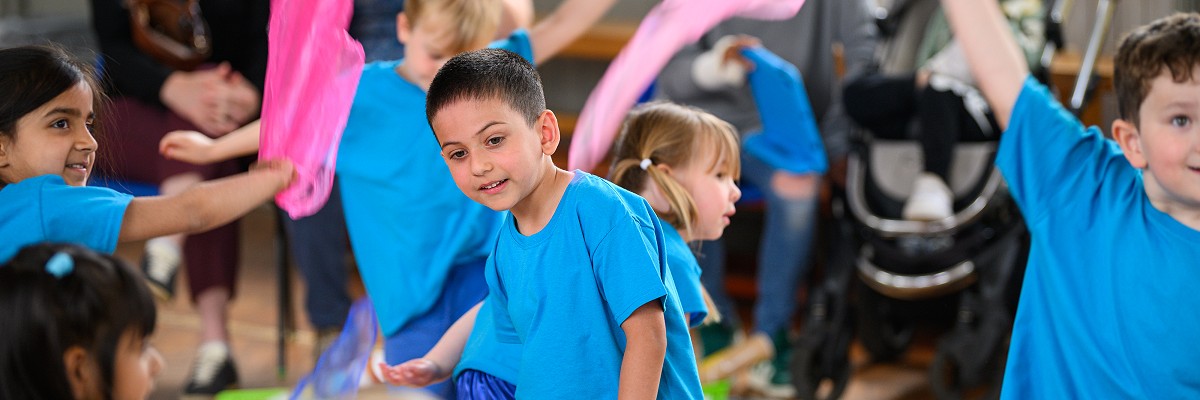 Reception children wearing blue t-shirts dance in a group, waving colourful scarves above their heads