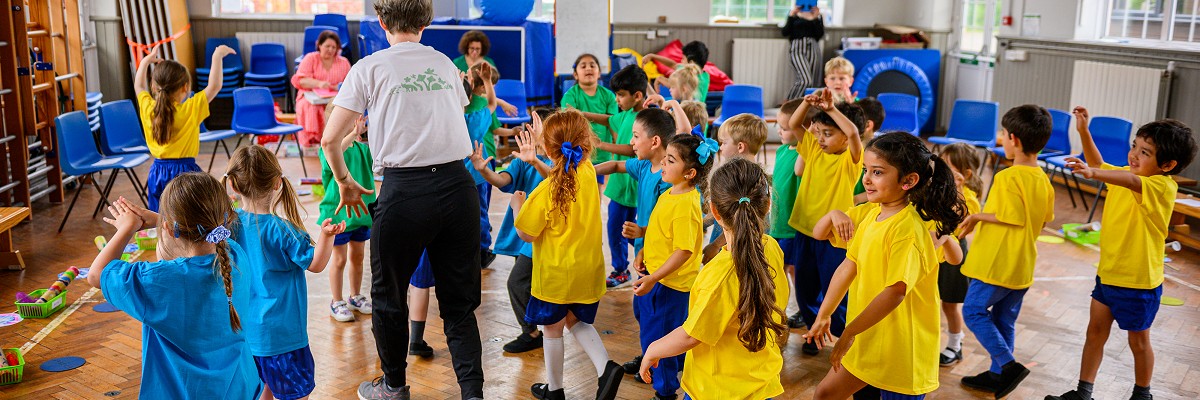 A class of reception children in brightly coloured t-shirts follow their dance teacher in a circle