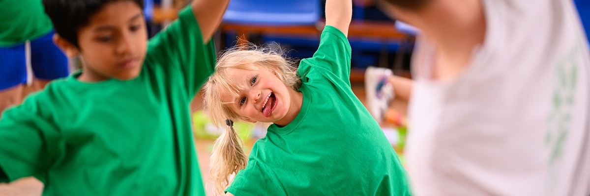 Children in green t-shirts follow a dance teacher, stretching over to the side