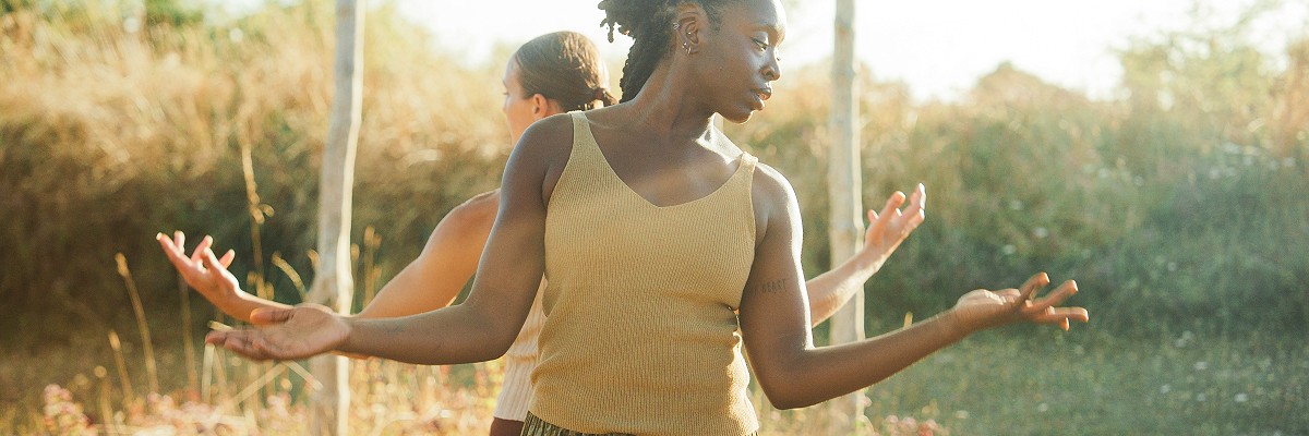 Two dancers stand back to back, their arms bent in at the elbows but with hands opening out to the sides. They stand in front of a series of wooden posts standing tall at the centre of a Bronze Age pond barrow.