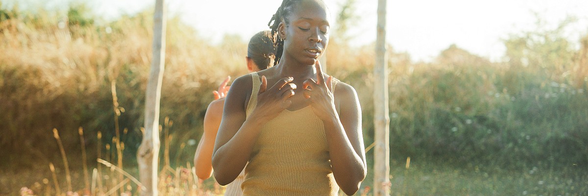 Two dancers stand back to back, their arms bent in at the elbows and their hands reaching towards their faces, caught in a moment of contemplation. They stand in front of a series of wooden posts standing tall at the centre of a Bronze Age pond barrow.