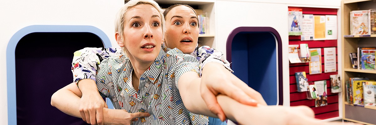 Two dancers dressed in bright colours perform in a library - they both point outwards into the camera, as if looking for something in the distance, about to start their adventure.