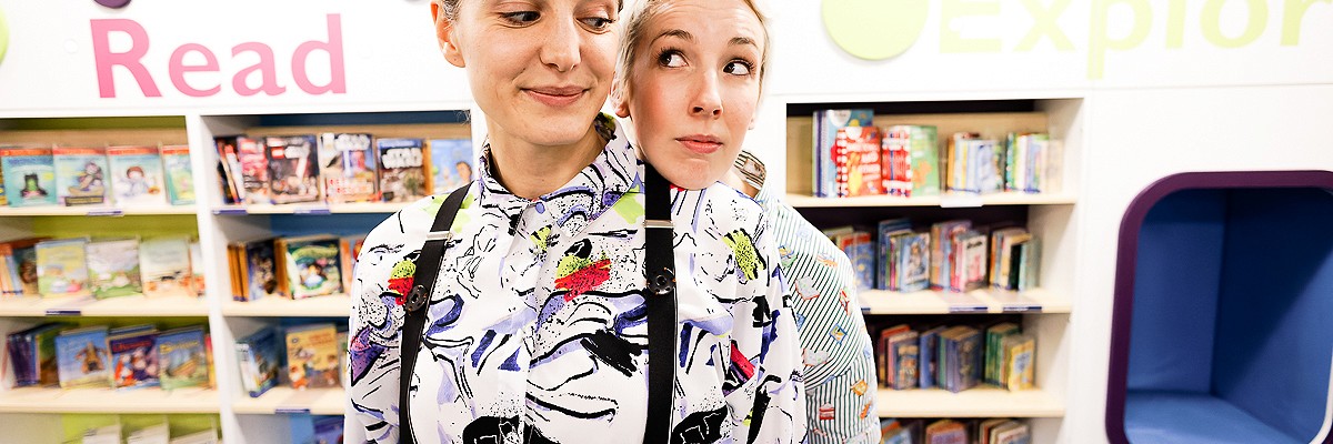 Two dancers, The Story Detectives, dressed with brightly coloured trousers and patterned shirts, stand in front of a wall of books in a library. Particular - the Story Detective in front - looks to her side as Playful, the Story Detective standing behind, rests her chin on Particular's shoulder. Playful looks very cheeky.