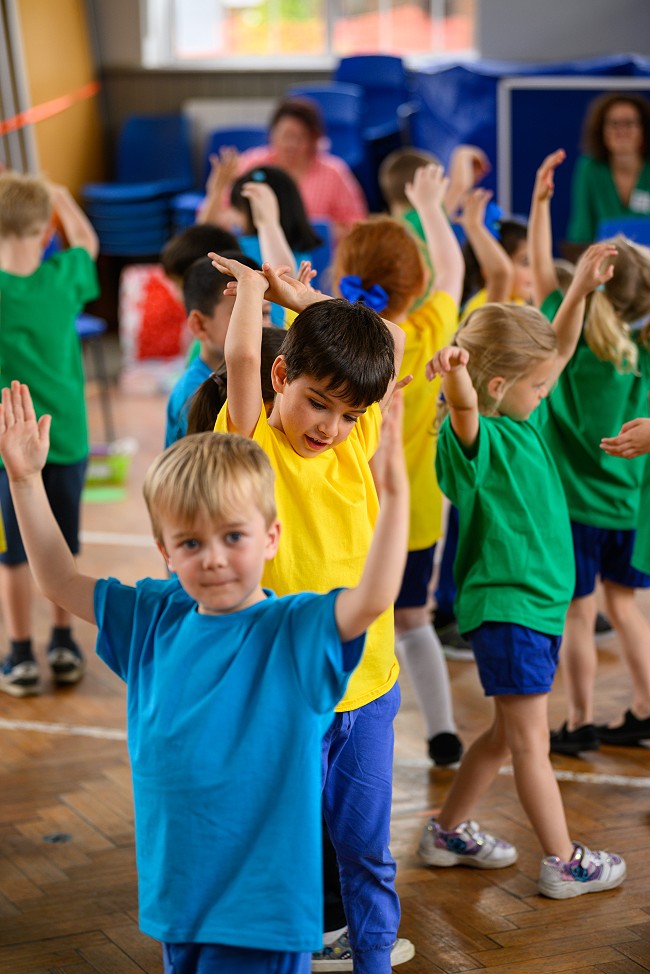 Primary school children with colourful blue, yellow and green t-shirts dance and reach their arms up to the sky
