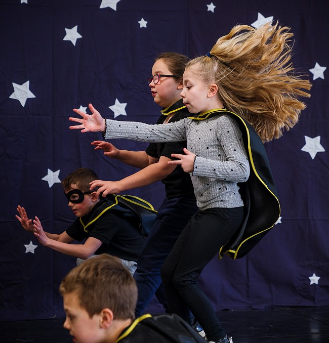 A group of primary school children perform their Villain dance in front of a blue backdrop covered with silver stars. The children all reach forward strongly with their arms, and they all wear black villain capes trimmed with yellow.